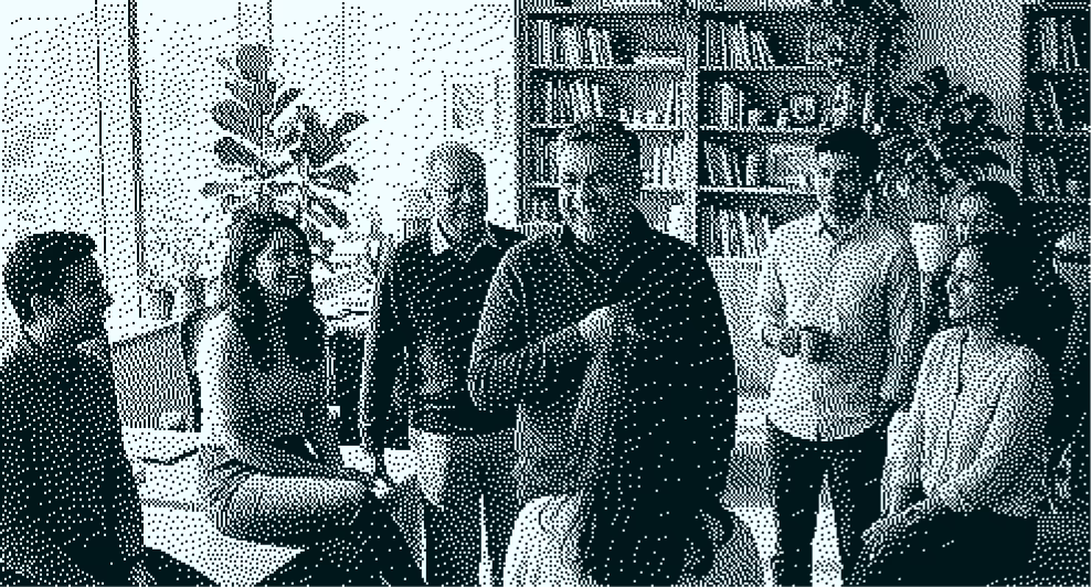 A group of seven people in a casual meeting in a room with bookshelves and plants.