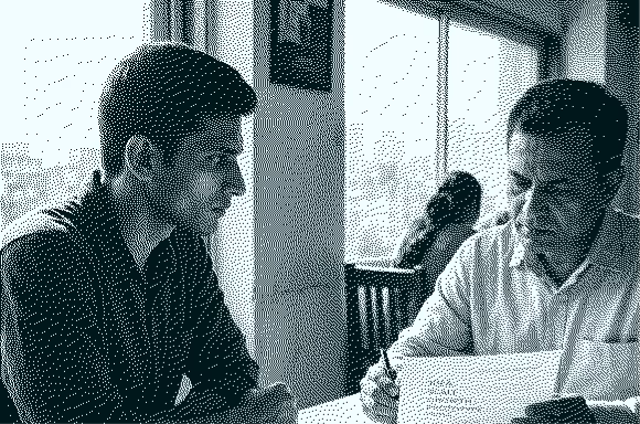 Two men in a cafe discuss paperwork with a window and another person in the background.