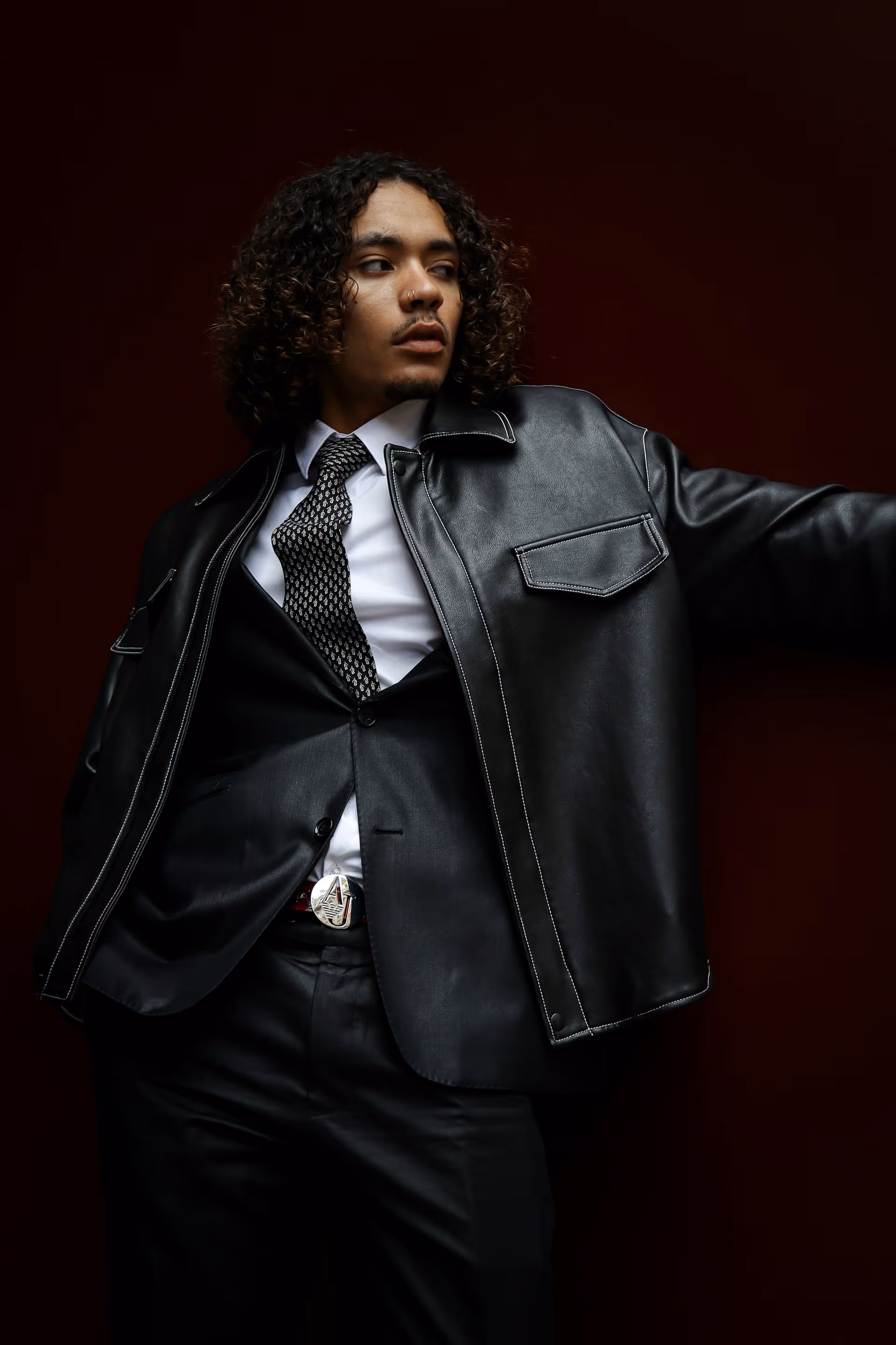 Man with curly hair wearing a black leather jacket, suit, patterned tie, and belt with a letter A buckle, against a dark background.