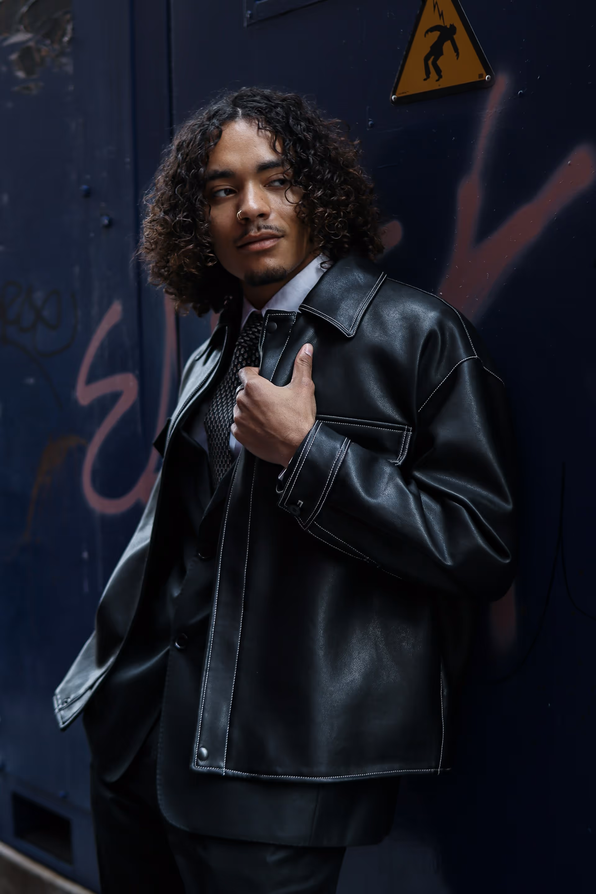 Man with curly hair wearing a black leather jacket and patterned tie leaning against a dark wall with graffiti.