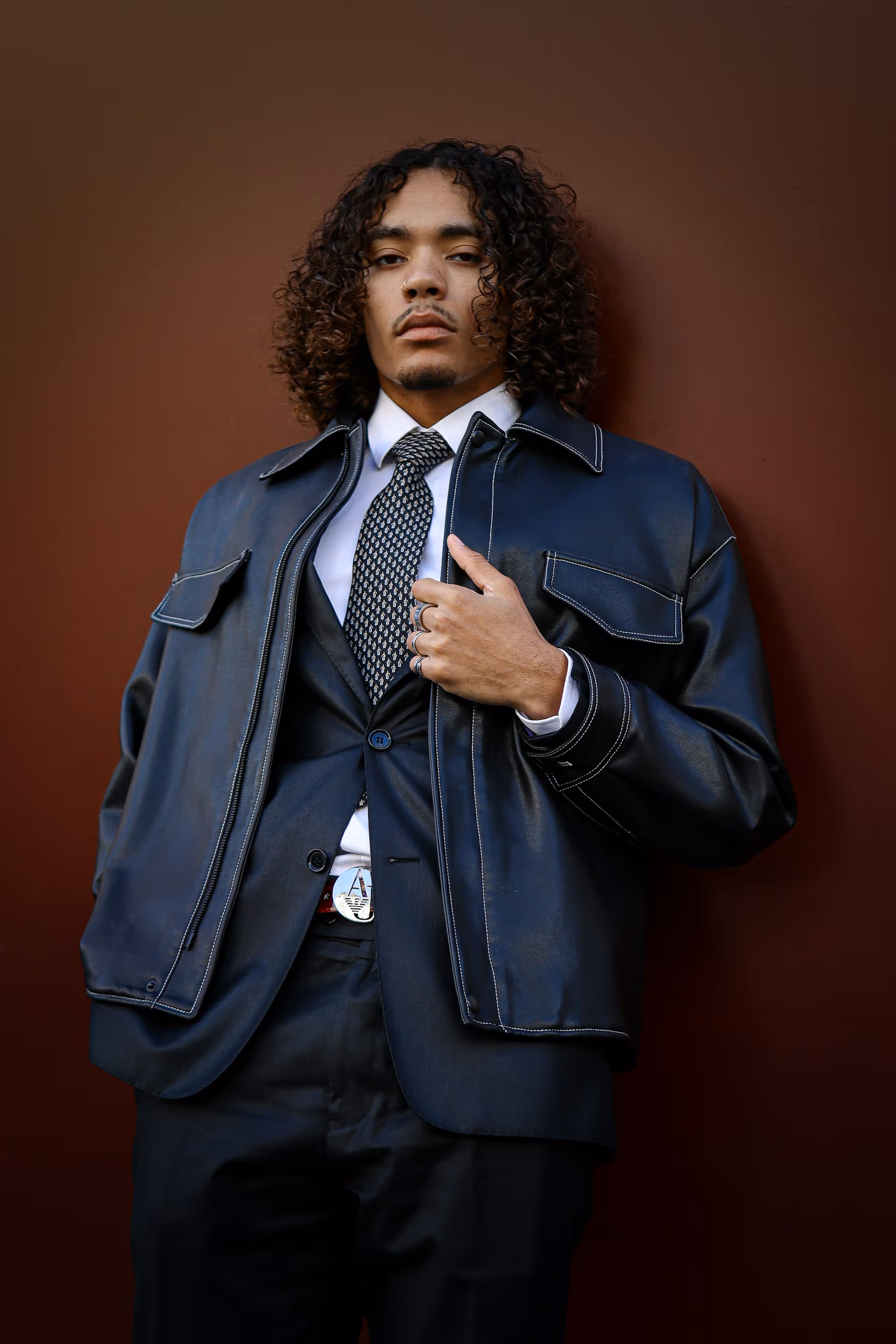 Man with curly hair wearing a black leather jacket over a suit, white shirt, patterned tie, and black pants, standing against a brown background.