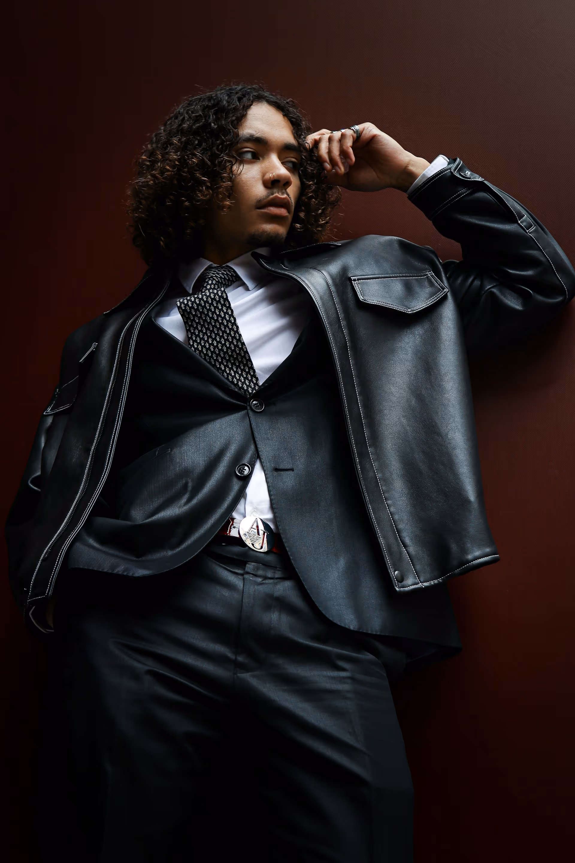 Man with curly hair wearing a black leather jacket over a black suit, white shirt, and patterned tie, posing against a dark background.