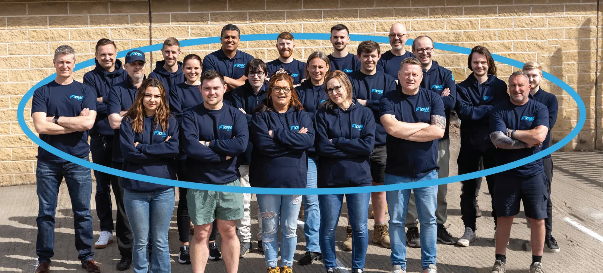 Group of adults wearing navy blue Flow branded shirts posing outdoors against a brick wall.