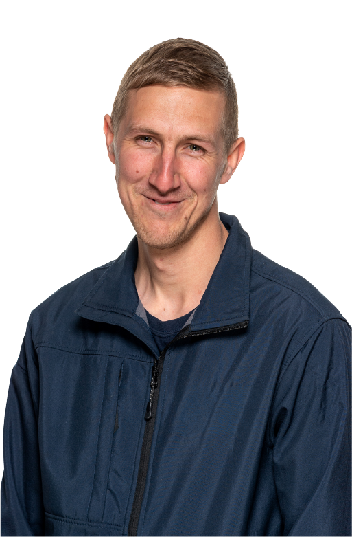 Smiling man with short light brown hair wearing a navy blue zip-up jacket over a dark shirt, standing against a white background.