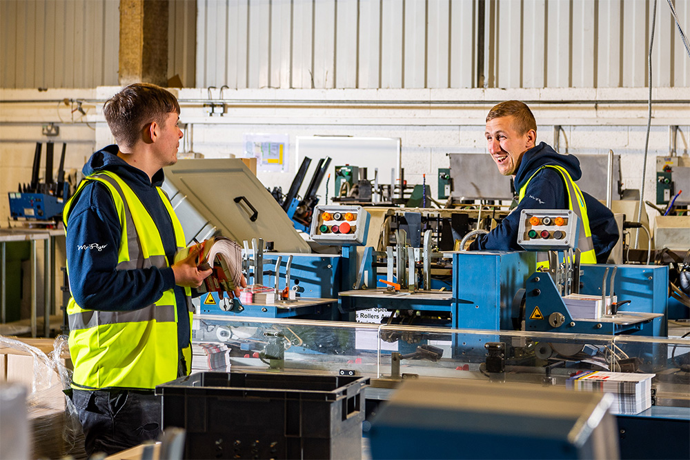 Two factory workers wearing high-visibility vests and dark hoodies operating a blue industrial machine in a brightly lit workshop, holding stacks of printed materials.