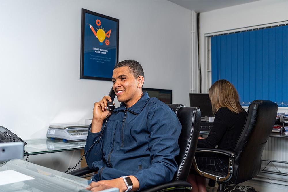 A man in a blue jacket sitting in an office chair and talking on a corded phone while smiling, with a woman working at a computer in the background.