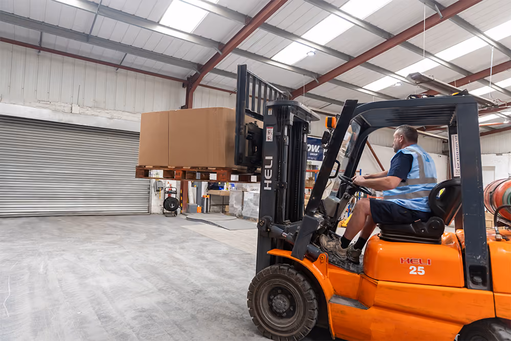 A man wearing a blue safety vest operates an orange HELI forklift lifting two large cardboard boxes on a wooden pallet inside a spacious warehouse with concrete floors and a metal roller door.
