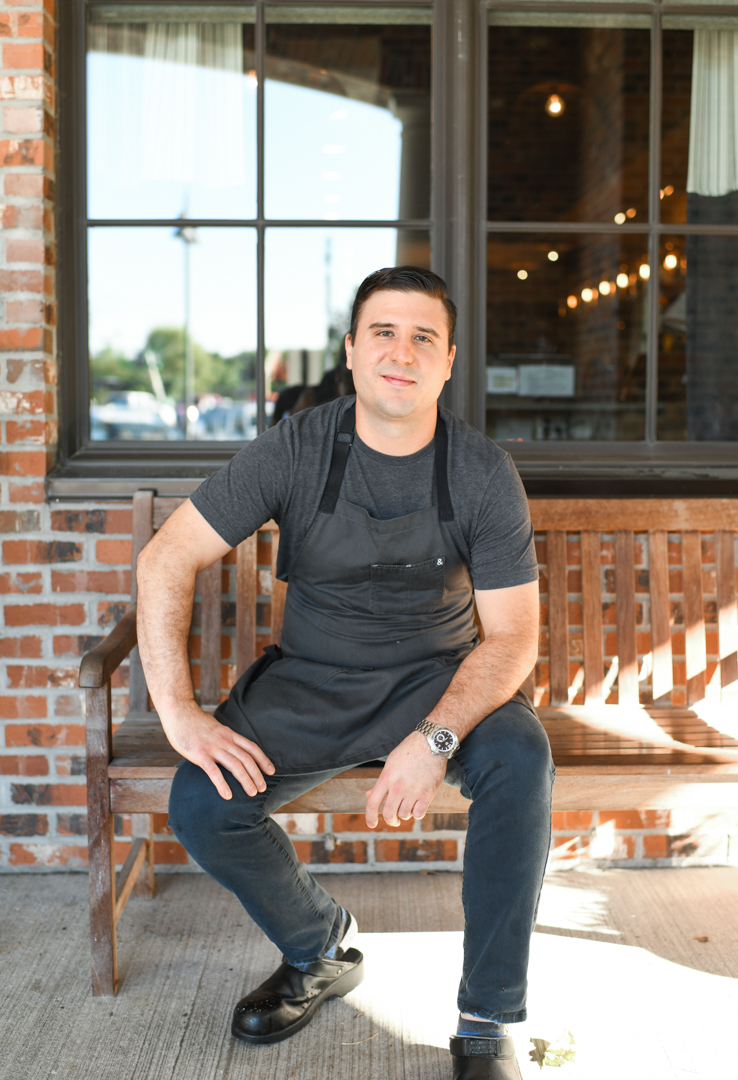 A man wearing a black apron and a gray shirt sitting on a bench.