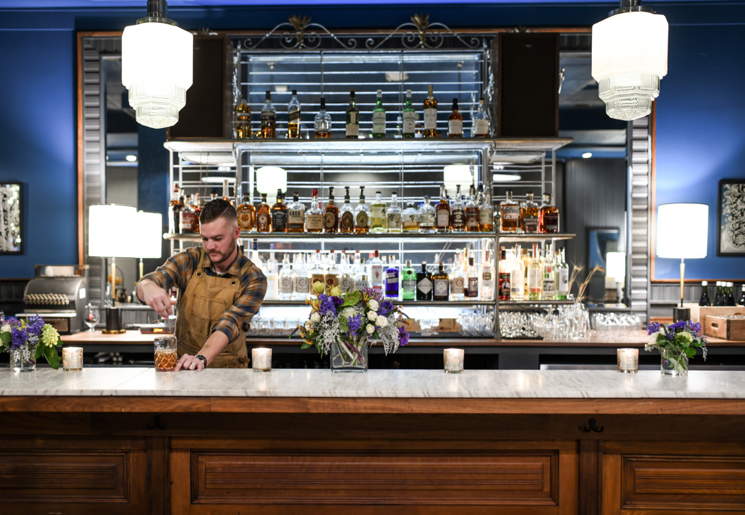 A man pouring a drink at a bar with a shelf full of liquor.