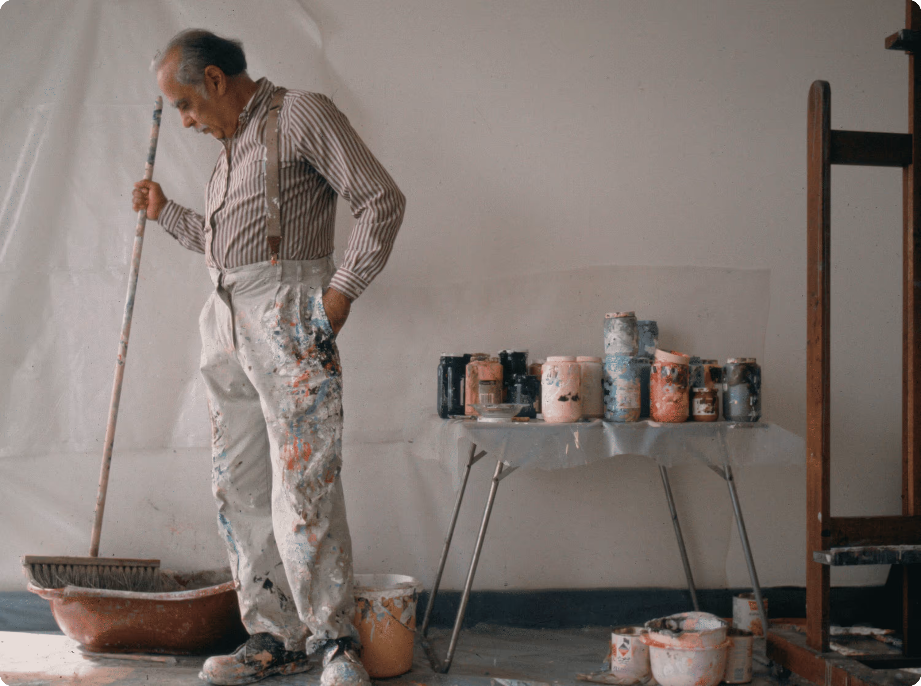 Ed Clark in paint-splattered overalls holding a broom and standing next to a table with many paint cans and supplies.