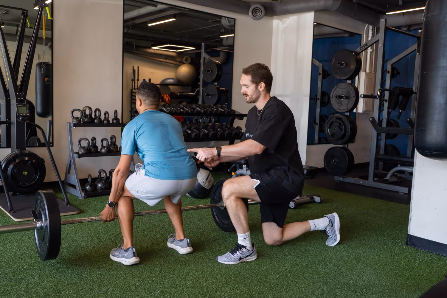 Trainer guiding man lifting barbell in gym with weights and kettlebells in background.