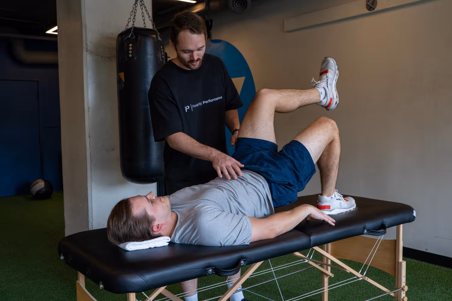 Trainer guiding a man lying on a table doing a leg raise exercise in a gym setting.