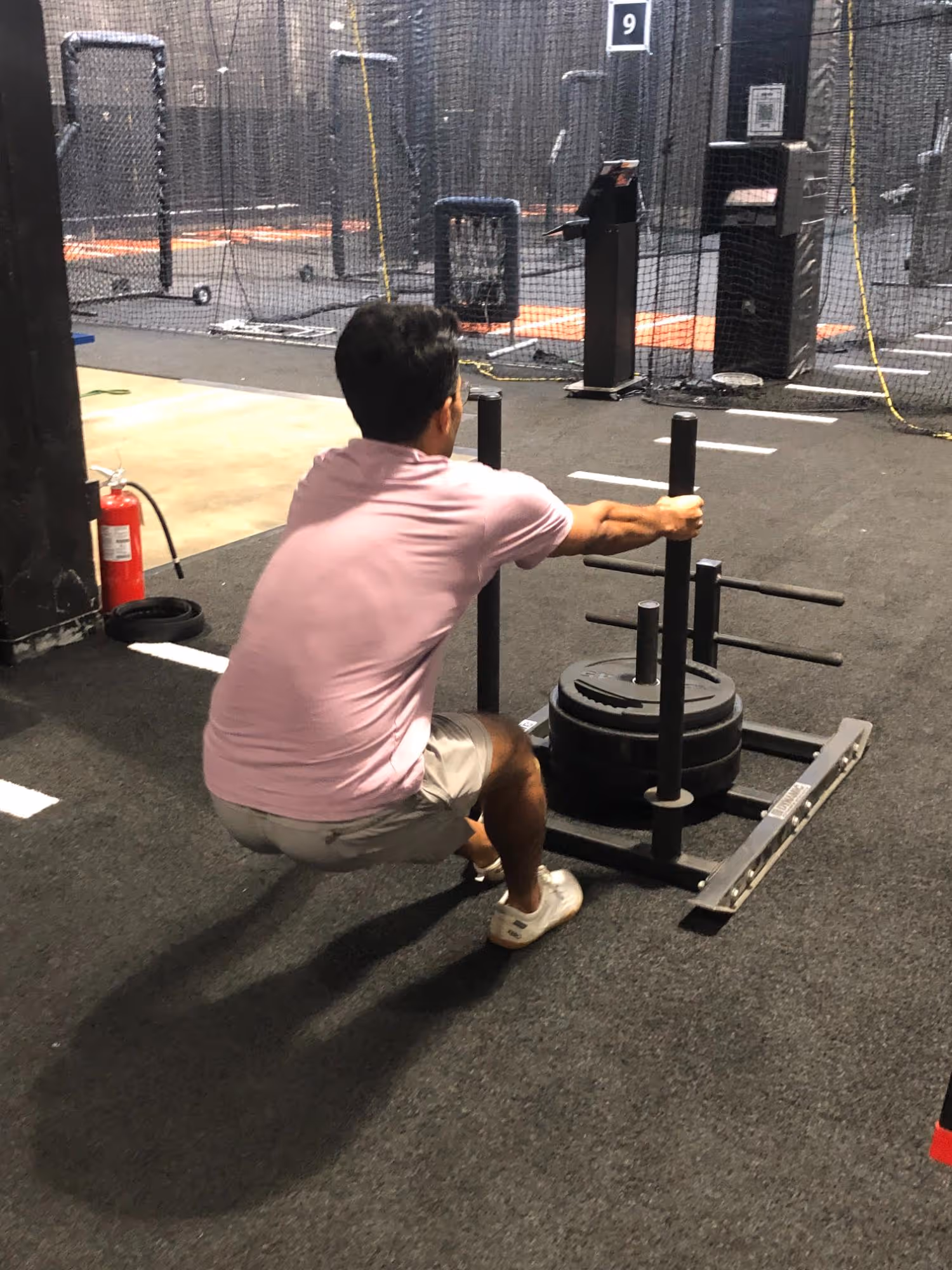 Man in pink shirt and shorts squatting while holding handles of weighted sled in gym.