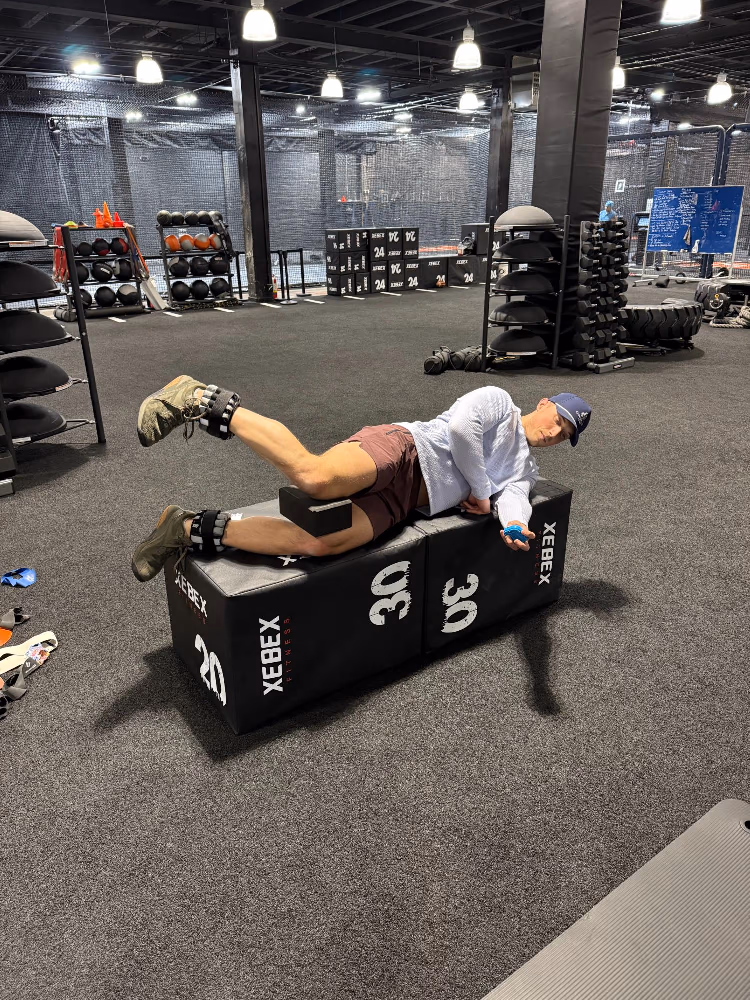 Man lying sideways on a padded block exercising leg lifts with ankle weights in a gym.