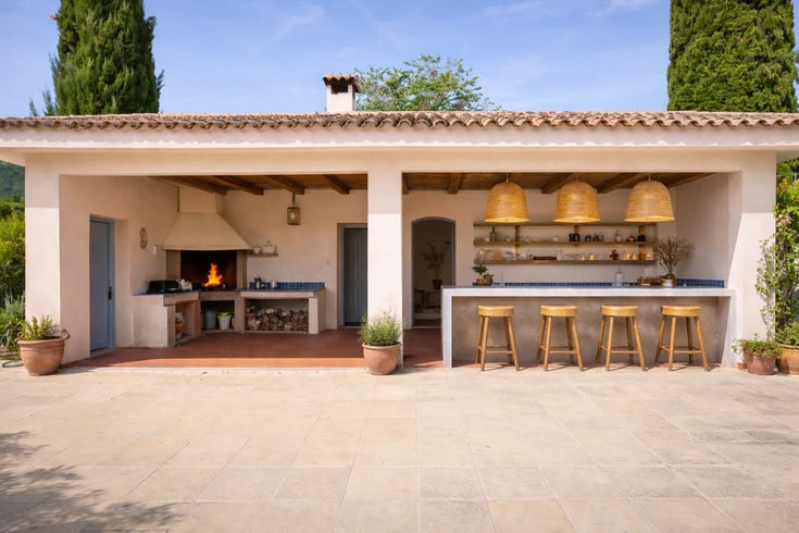 Cuisine extérieure avec un barbecue allumé, un comptoir avec quatre tabourets en bois et des suspensions en osier sous un toit à tuiles.