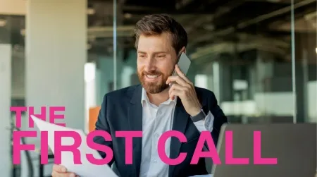 A smiling businessman in a dark suit talking on his mobile phone while reviewing printed campaign data, overlayed with the pink text "THE FIRST CALL"