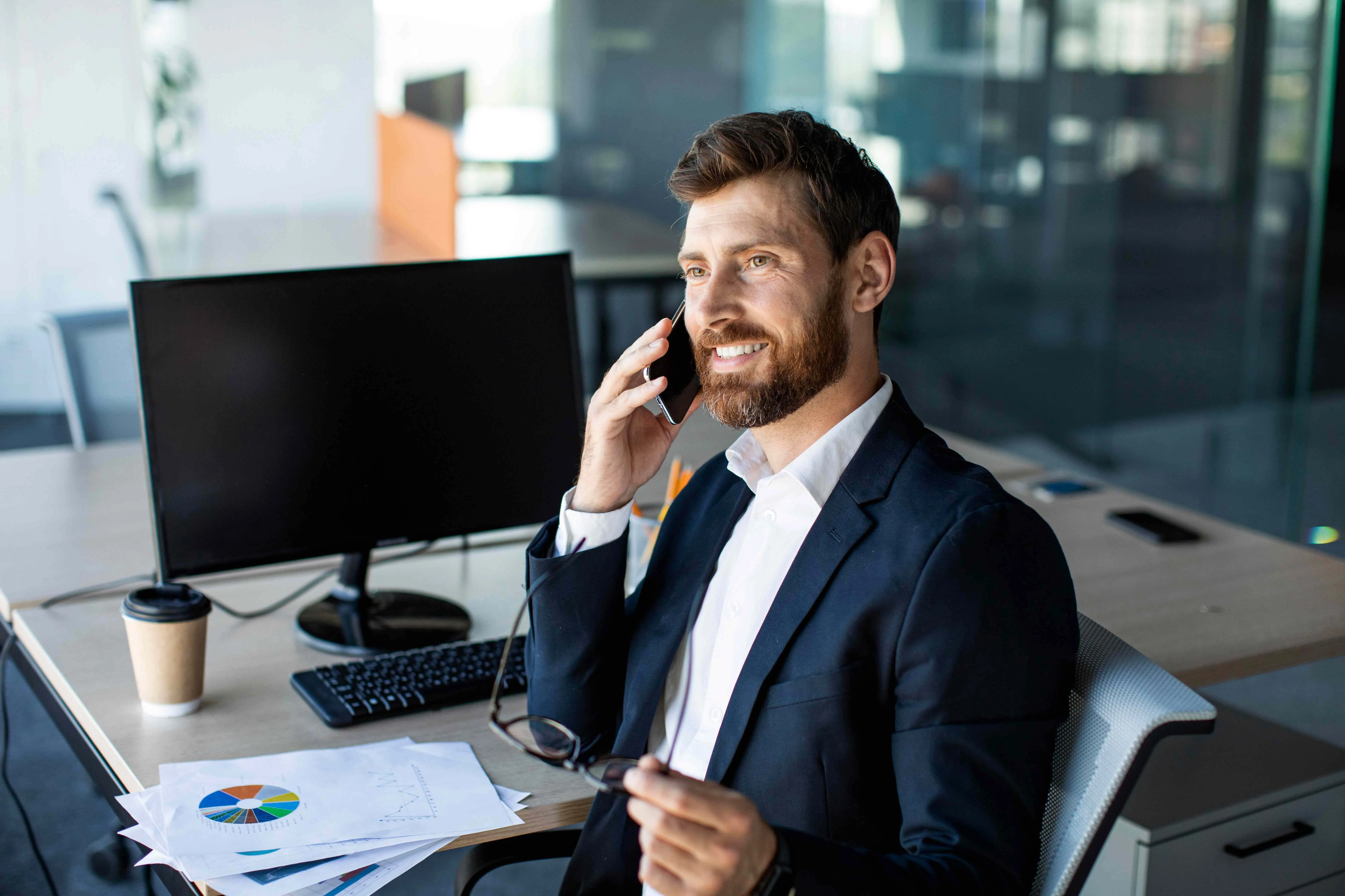 A smiling businessman in a navy suit sitting at an office desk, talking on a smartphone while looking at printed data charts, illustrating a successful marketing lead.