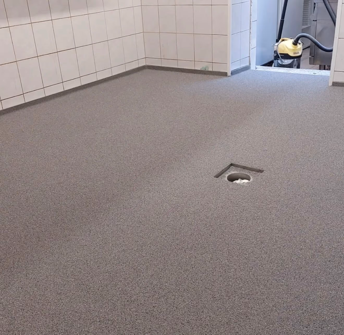 Zimmer mit grauem, nahtlosem Bodenbelag und weißen Wandfliesen mit einem Abfluss in der Mitte; hinten ein offener Raum mit einem gelben Staubsauger.