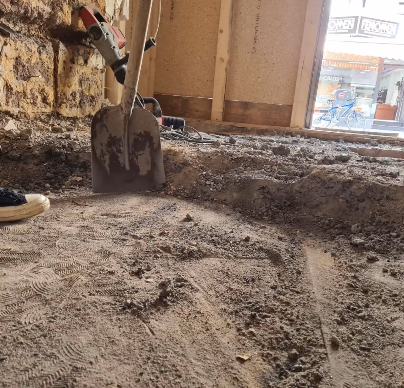 Eine Schaufel steckt im Boden in einem Raum mit schmutzigem, unebenem Boden, umgeben von einer Steinwand, Bauholz und einem Fenster zur Straße, auf der ein blaues Fahrrad steht. Boden wurde zurückgebaut.