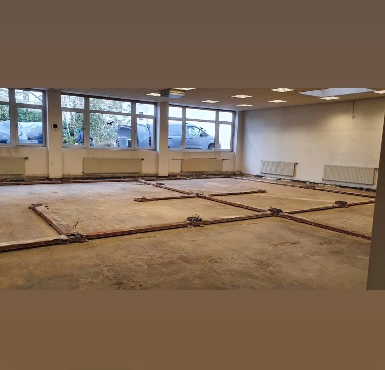 Leerer Raum mit nacktem Betonboden und sichtbaren Metallrahmen auf dem Boden, große Fenster und Heizkörper an den Wänden, draußen sind Fahrzeuge zu sehen.