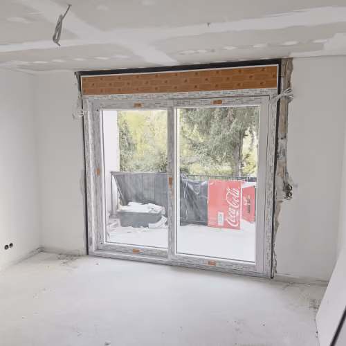 Unfertiges Zimmer mit weiß verputzten Wänden und einer großen Doppelschiebetür, die auf einen Balkon mit Blick auf grüne Bäume. Neuer Estrich als Bodenbelag.