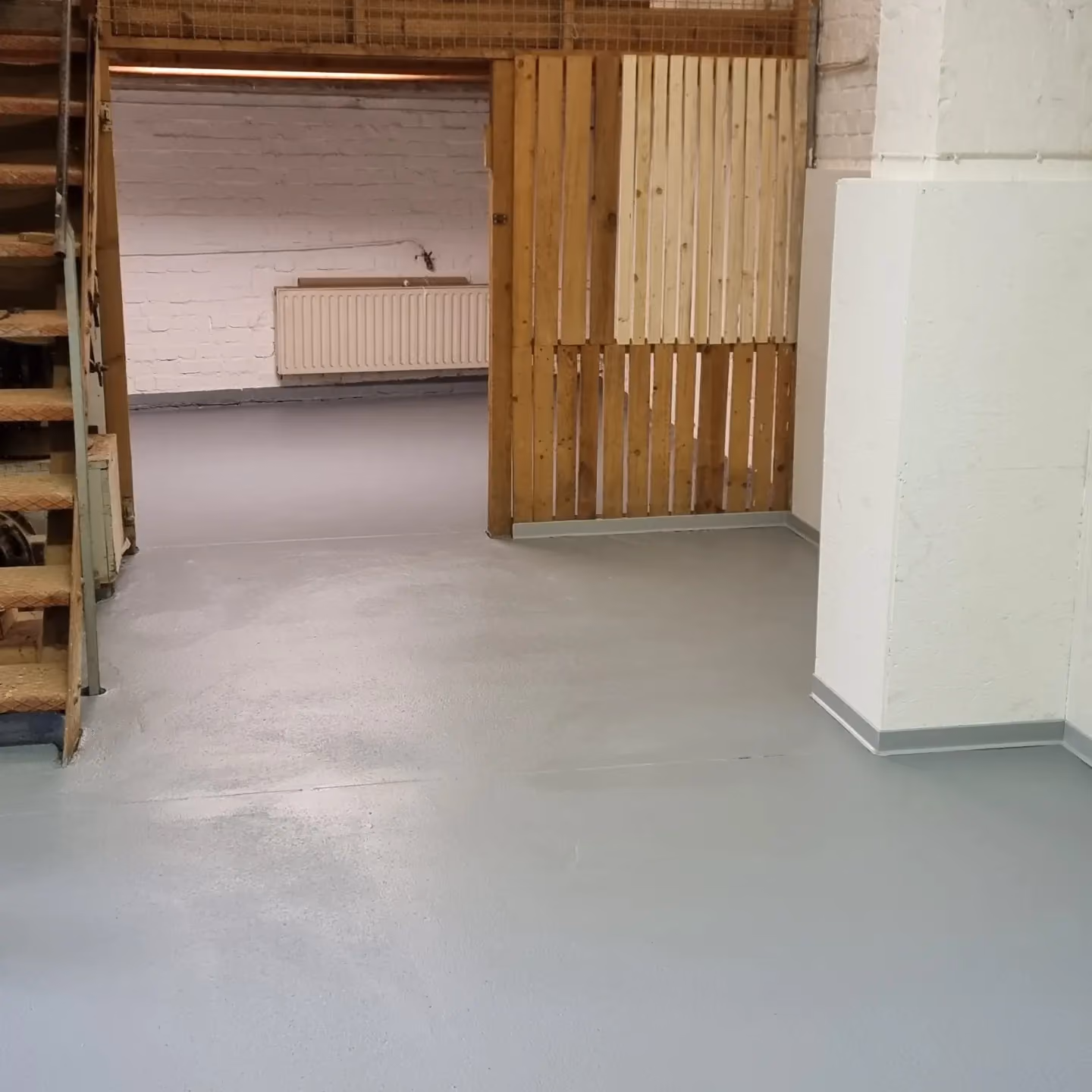 Heller, leerer Raum mit grauem Boden, weißen Wänden, einer Heizkörper mit Rohrinstallation und einer halb geöffneten Holztür rechts neben einer mit Holz verkleideten Wand.