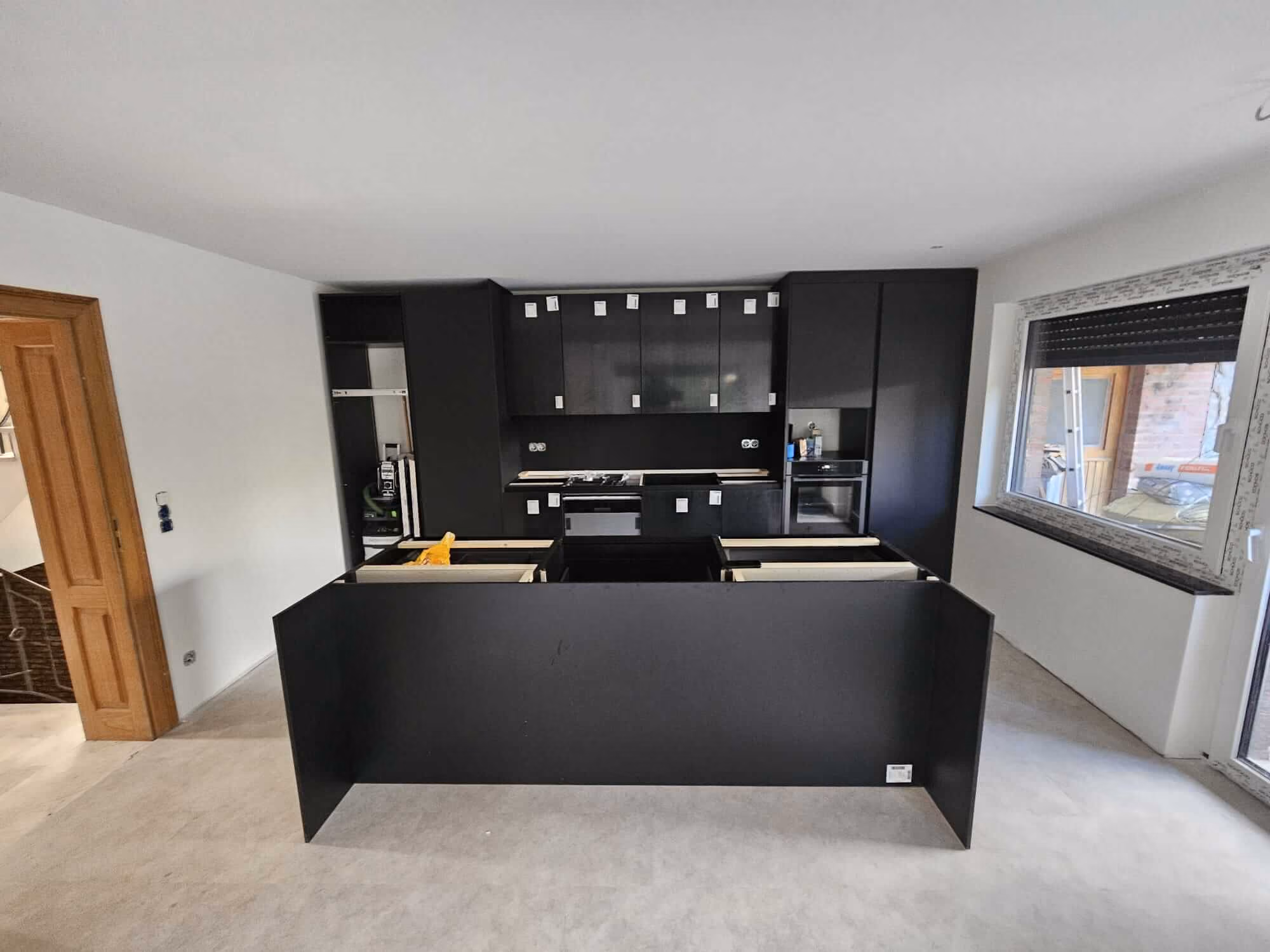 Moderne Küche im Umbau mit schwarzen Schränken und Arbeitsflächen, eingebautem Backofen, Fenster rechts und Holztür links. Moderner Bodenbelag in Hellgrau.
