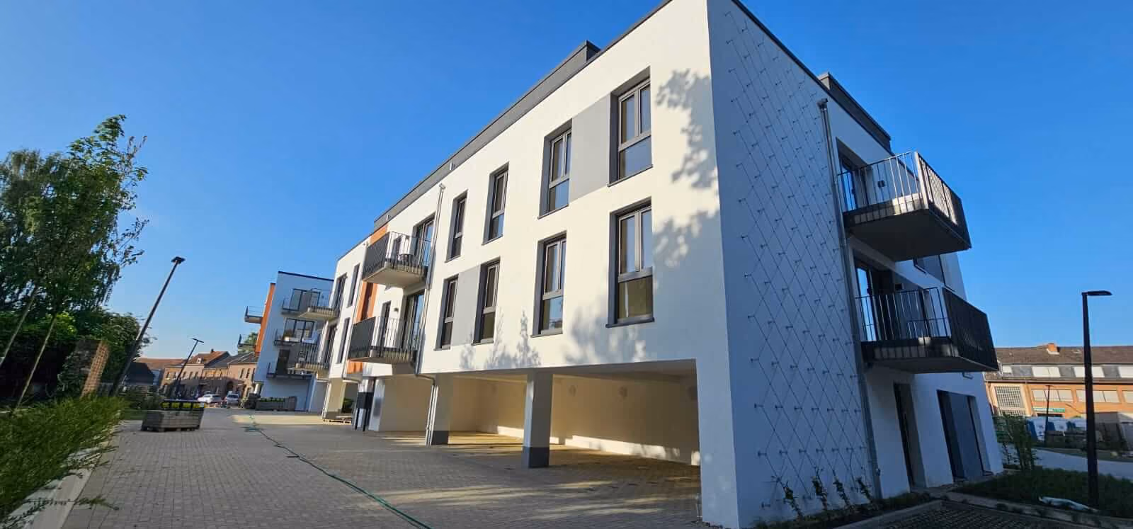 Moderner, weißer dreistöckiger Wohnblock mit Balkonen, Parkmöglichkeiten darunter, an sonnigem Tag mit blauem Himmel.
