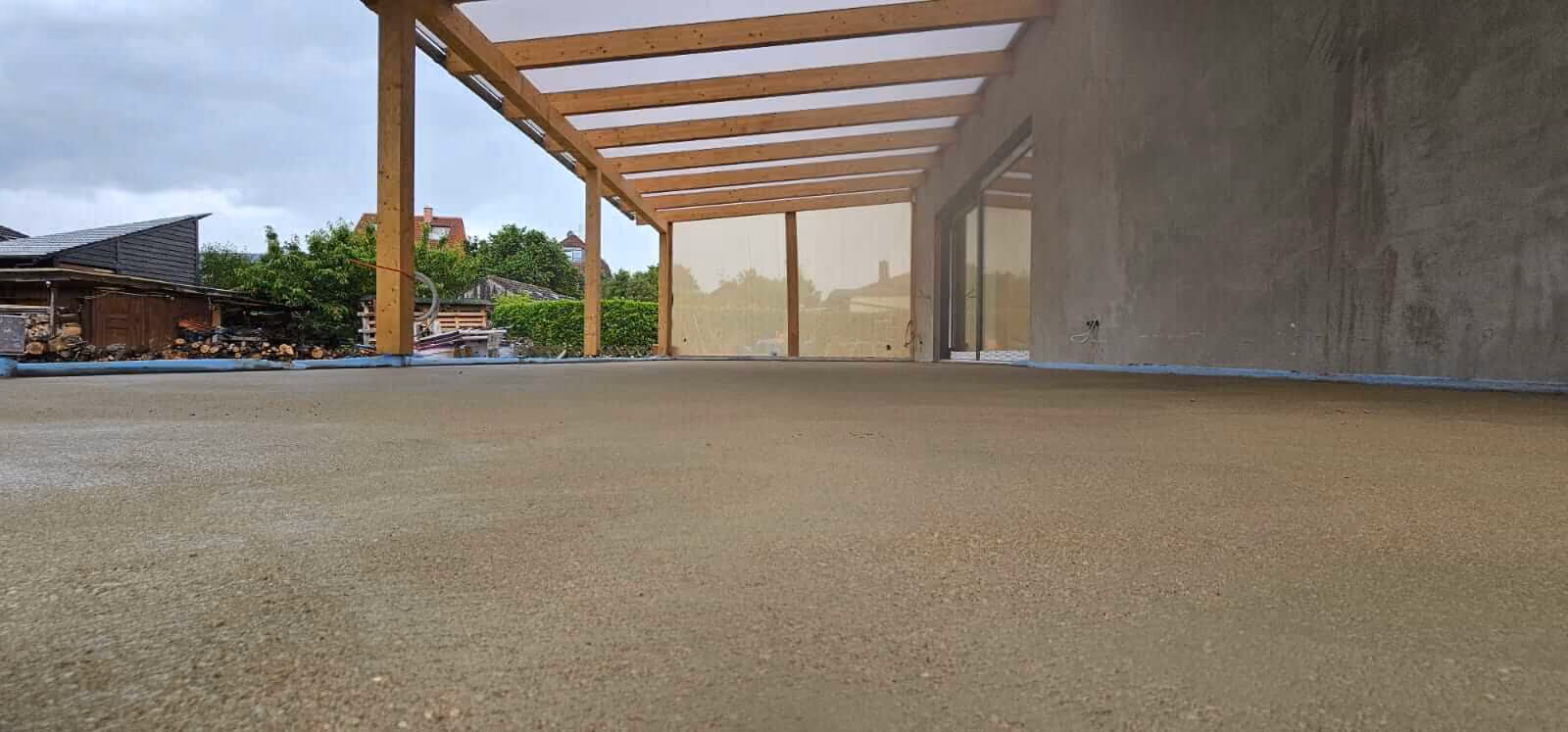 Blick über eine glatte, frisch gegossene Betonbodenfläche unter einem Holzrahmen-Dach mit angrenzender Betonwand und großen Fenstern an einem bewölkten Tag.
