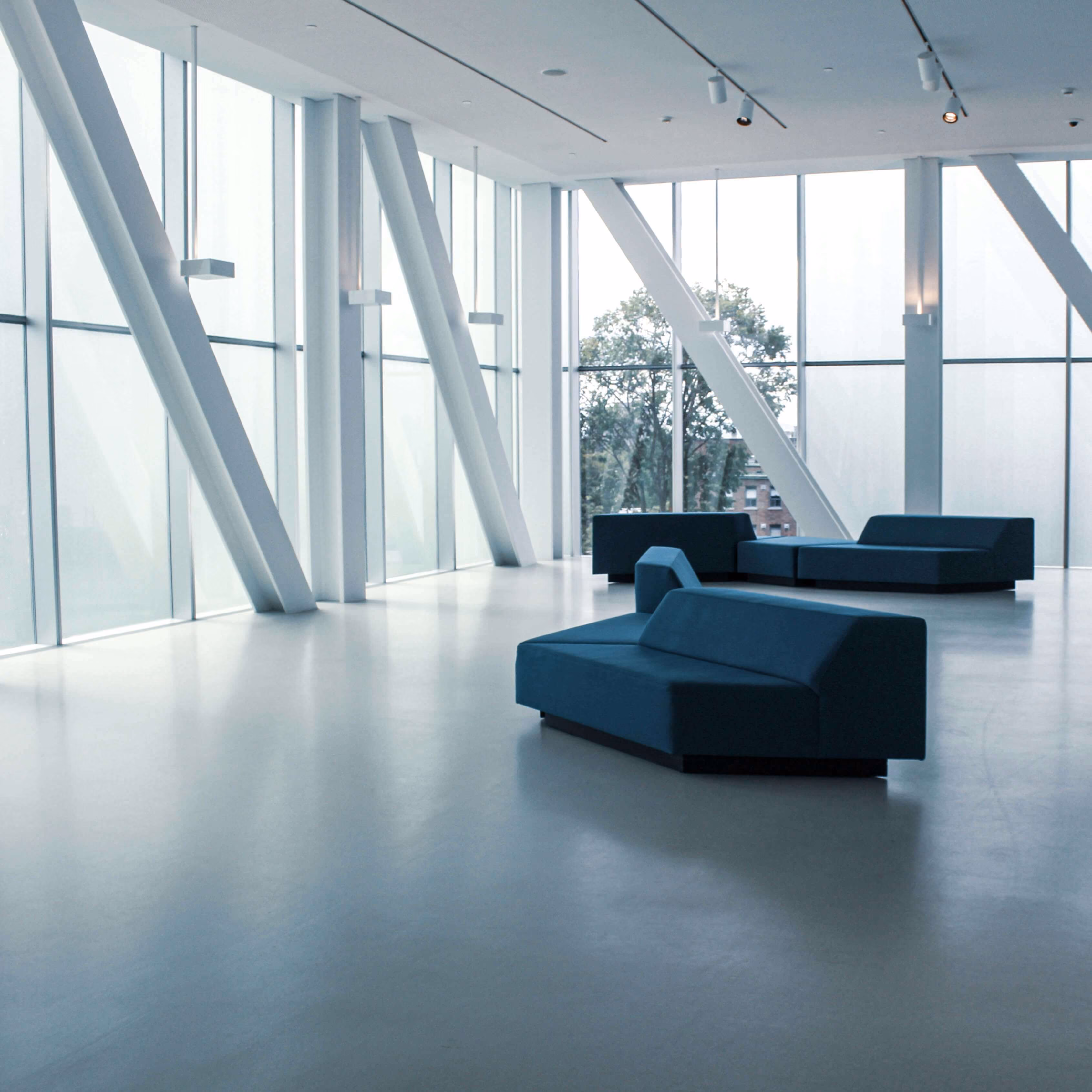 Heller, moderner Innenraum mit großen Fensterfronten, weißen Stützpfeilern und mehreren blauen Sofas auf einem glatten Boden.