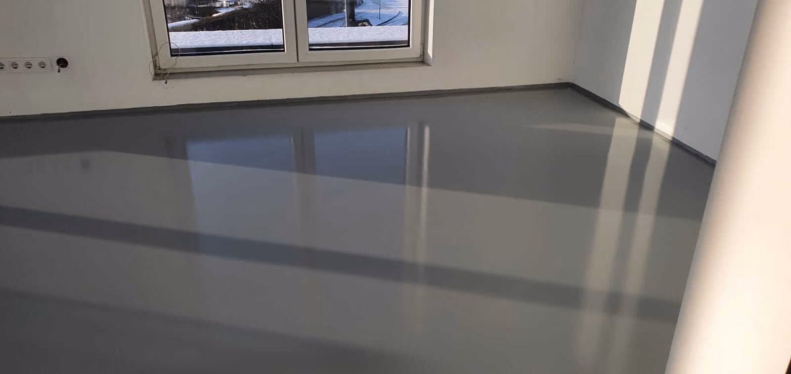 Leerer Raum mit glänzend poliertem grauem Boden, weiß gestrichenen Wänden und einem Fenster, durch das Tageslicht und Schatten hereinfallen.