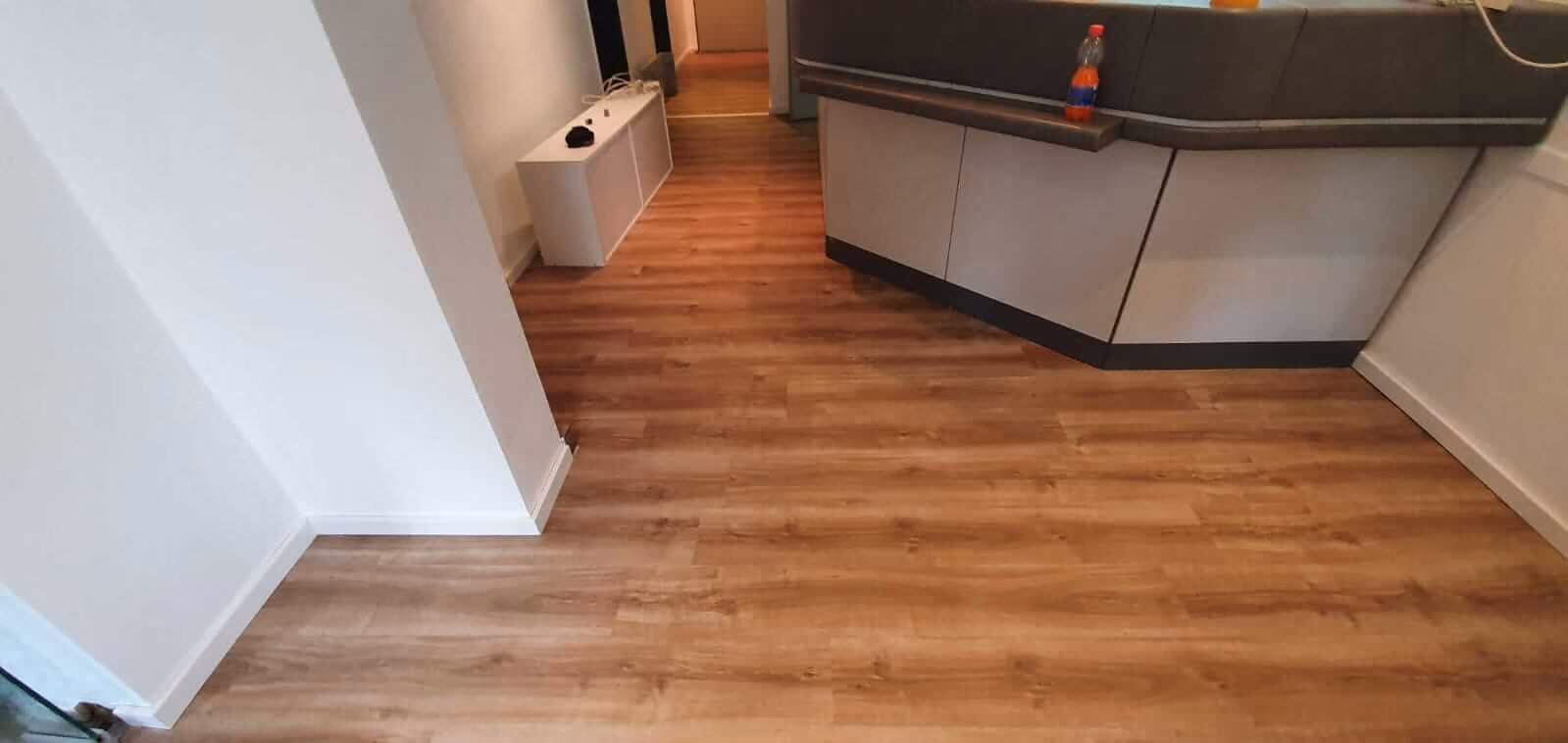 Raum mit braunem Holzboden, weiß gestrichenen Wänden, einem weißen Schrank gegen die linke Wand und einer grauen Theke rechts mit einer Flasche darauf.