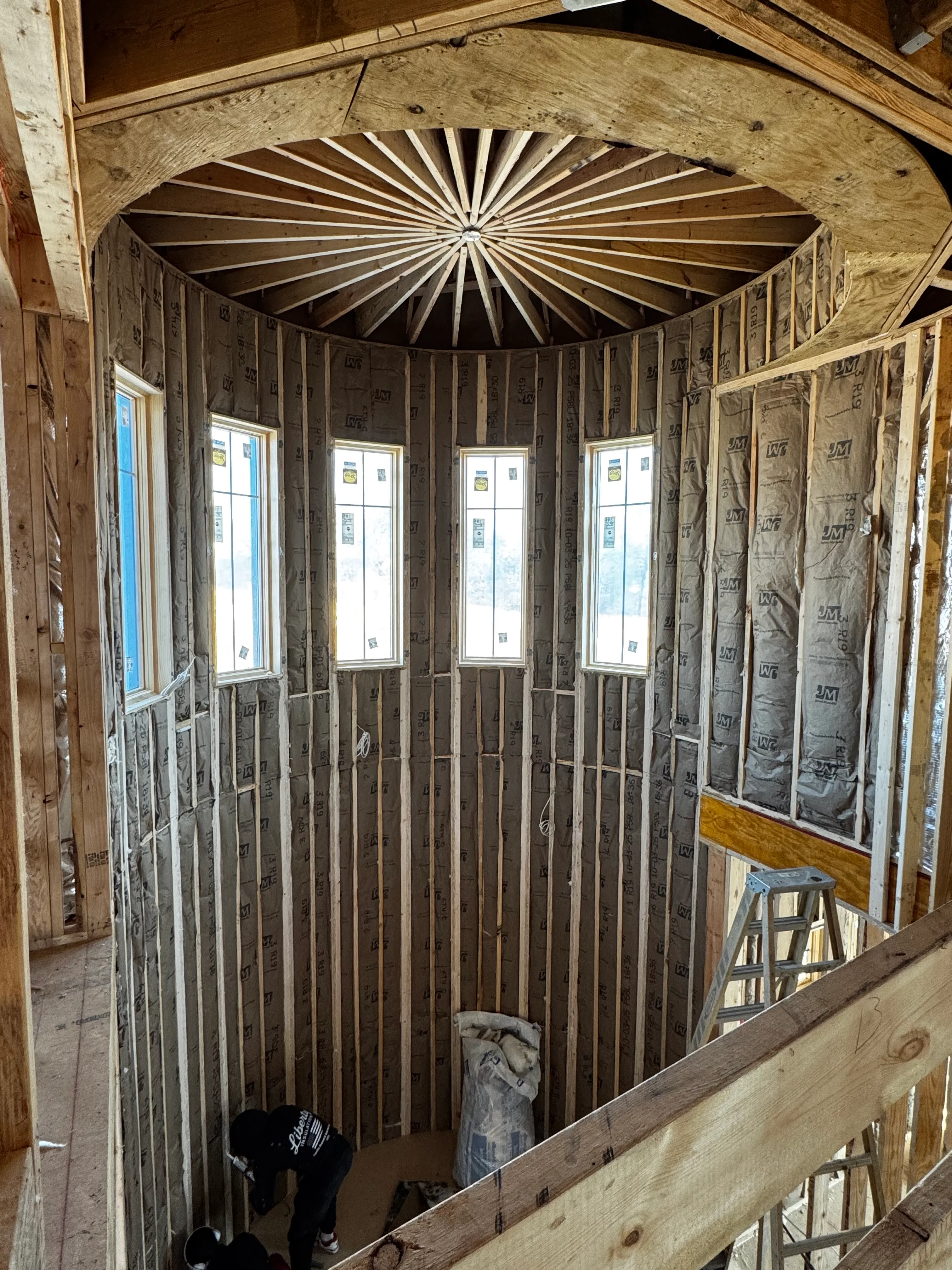 Interior of a circular wooden structure under construction with five tall narrow windows and insulation.