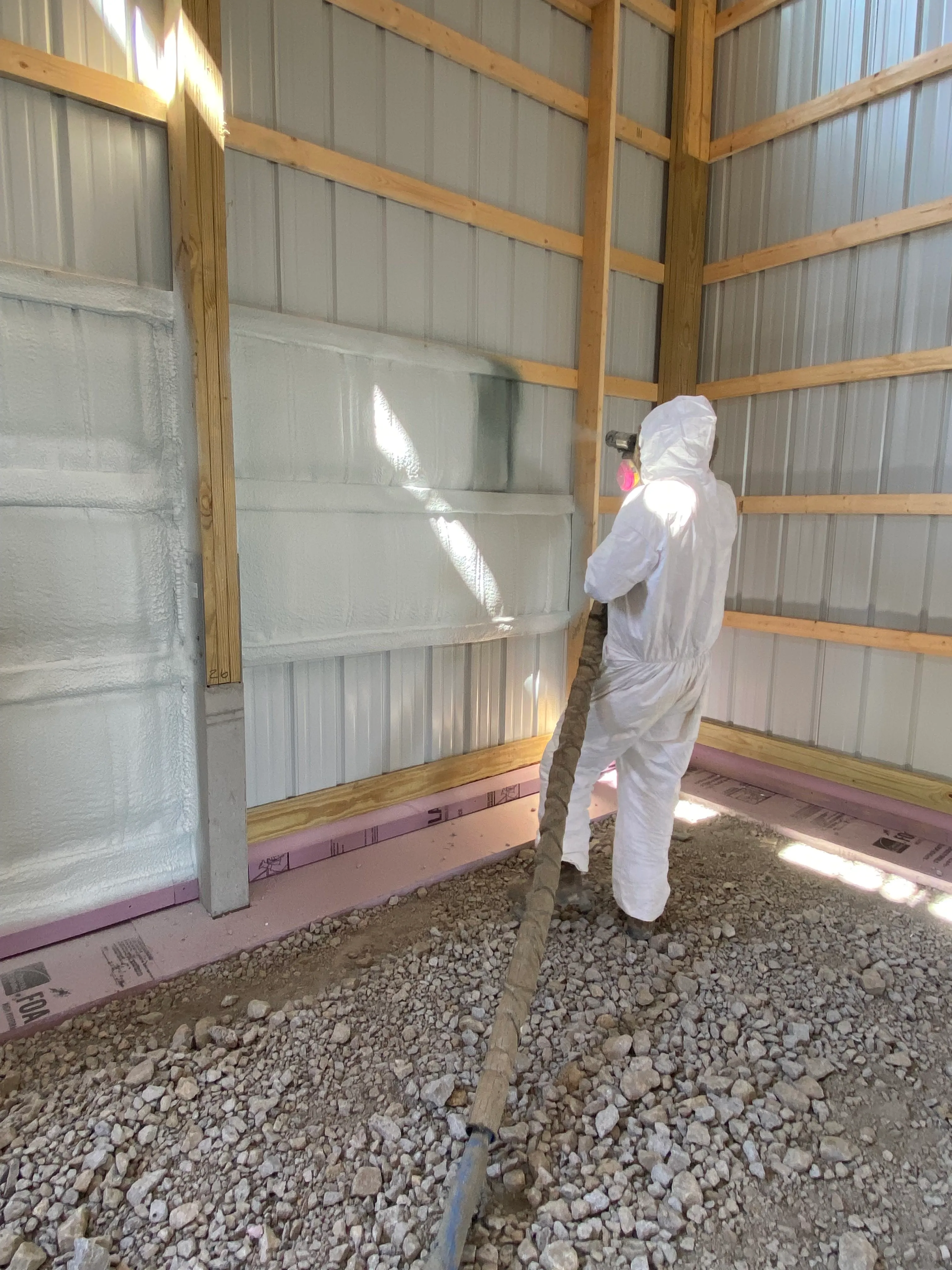 Worker in white protective suit spraying foam insulation on metal wall inside a building under construction.