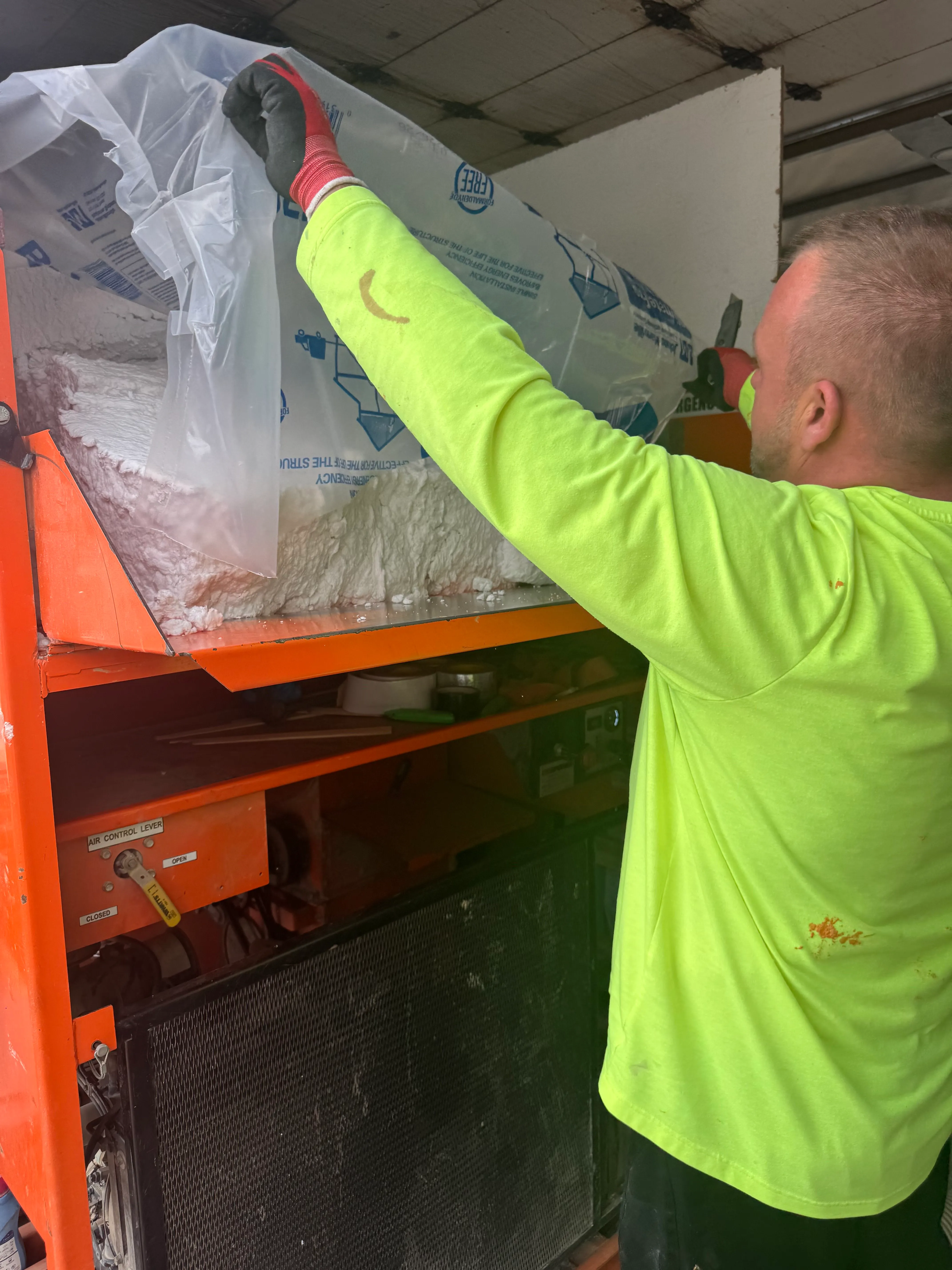 Worker in neon yellow shirt pours white insulation from plastic bag into large orange machine.
