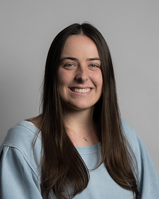 Smiling woman with long dark hair wearing a light blue sweater against a gray background.