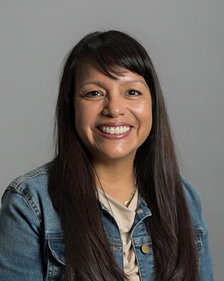 Smiling woman with long dark hair wearing a denim jacket and beige top against a gray background.