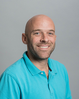 Smiling bald man wearing a turquoise polo shirt with a light gray background.