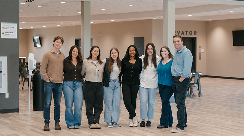 Group of eight casually dressed people standing indoors in a spacious room smiling at the camera.