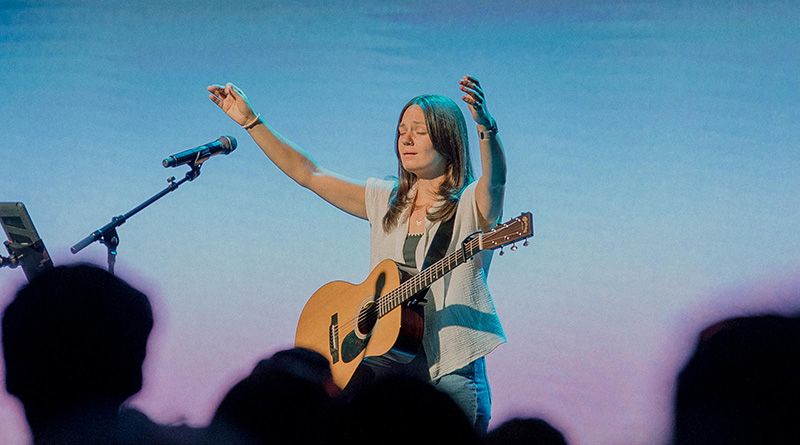 Woman with guitar raising her arms and eyes closed while performing on stage.