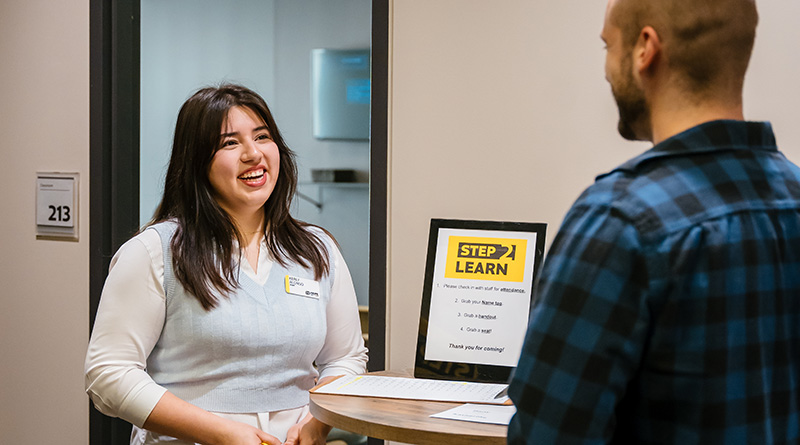 Smiling woman at a welcome desk talking to a man in a blue plaid shirt near a Step 1 Learn sign.