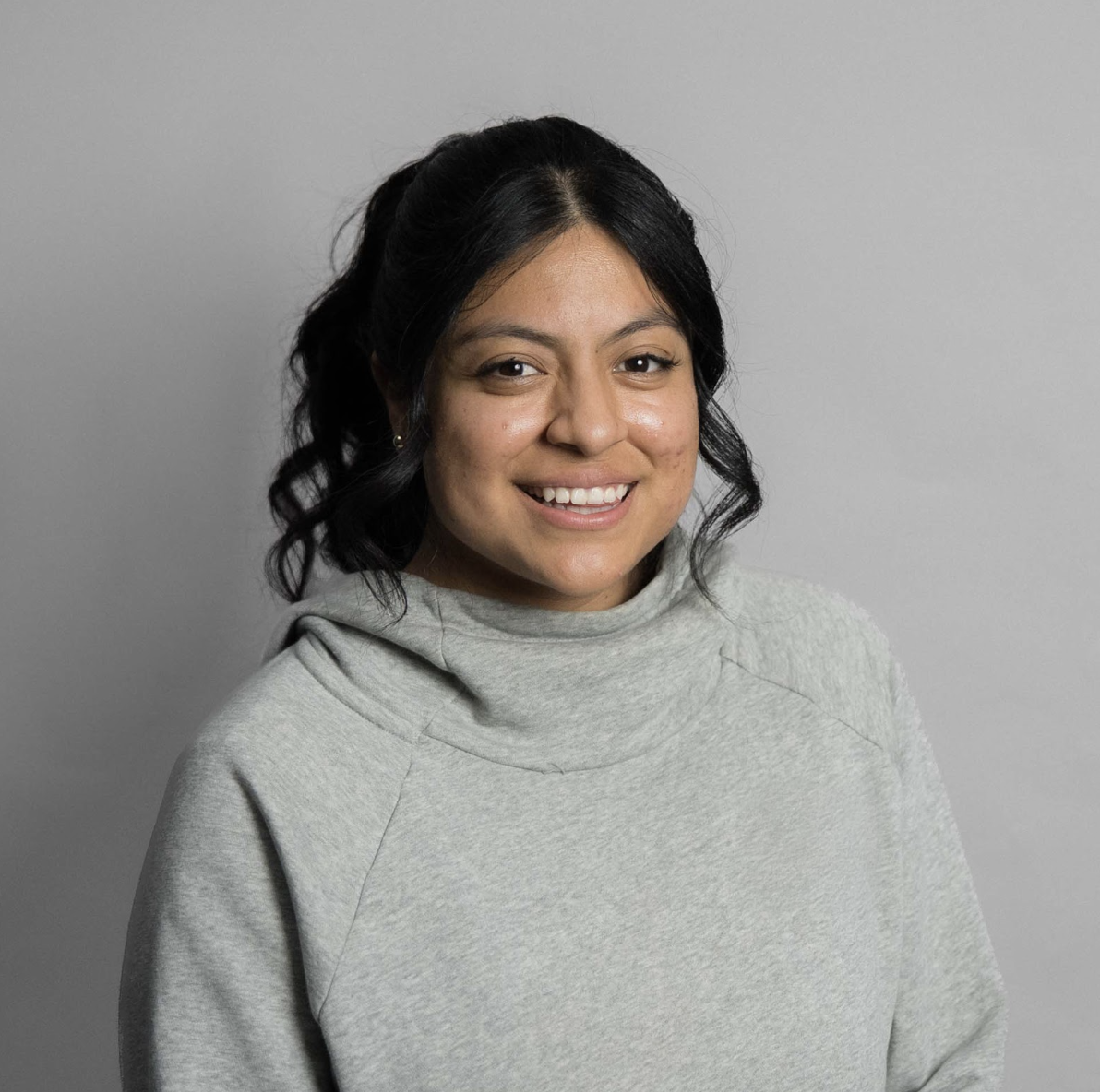 Smiling woman with long dark hair wearing a light blue sweater against a gray background.