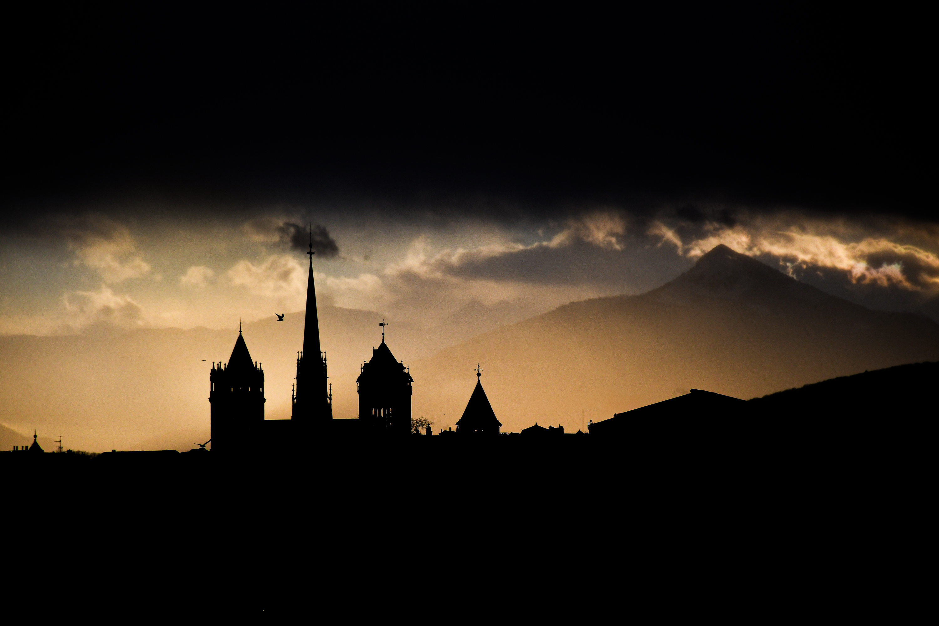 Silhouetted church towers and buildings against a sunset sky with mountains in the background.