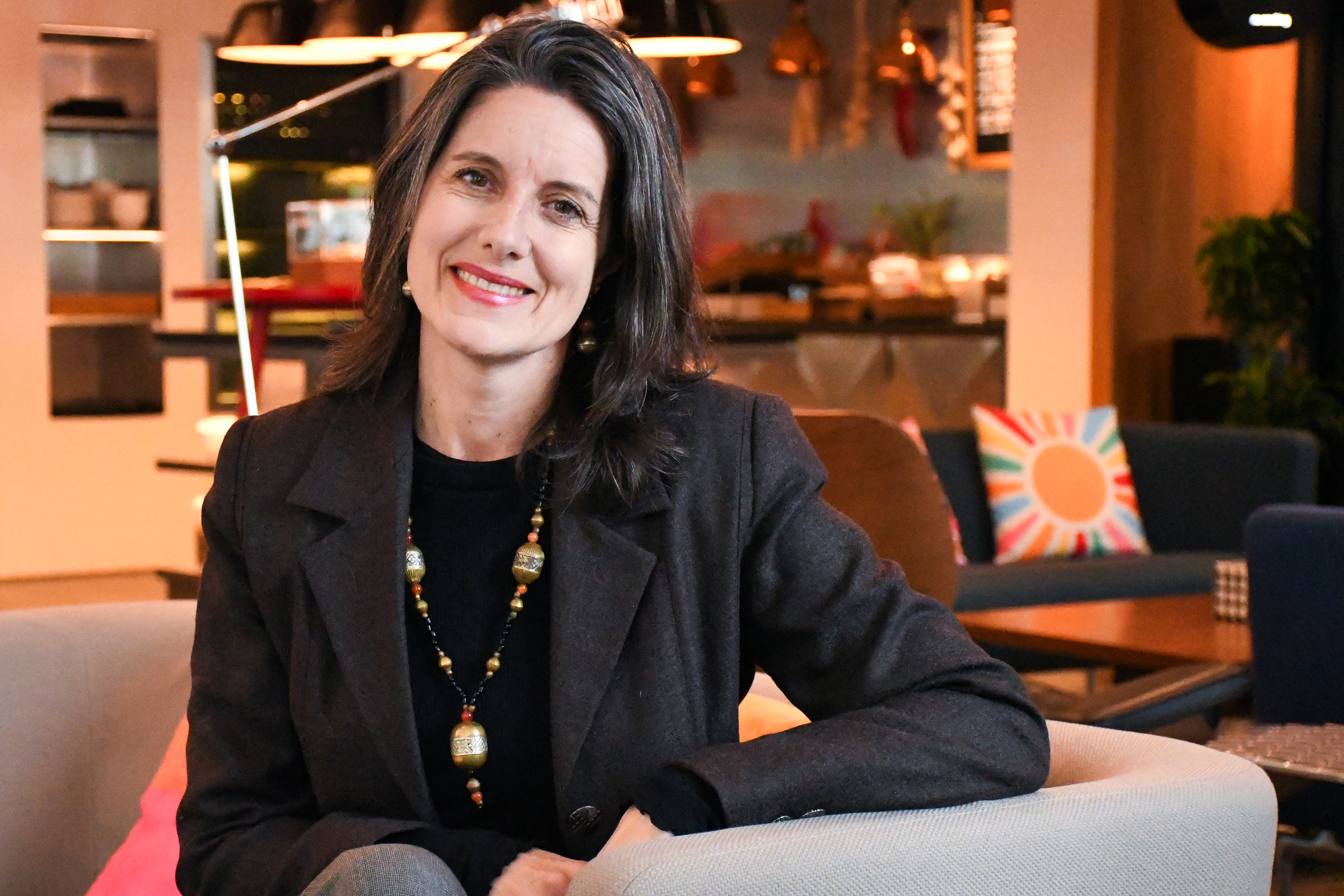 Smiling woman with dark hair sitting in a light-colored armchair in a cozy, warmly lit hotel lobby.