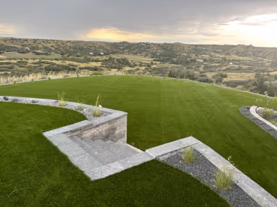 Aerial view of a green lawn bordered by stone steps and retaining walls with small plants, overlooking a wide valley under a cloudy sky.