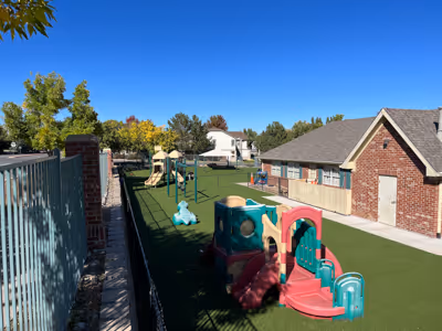 Outdoor playground area with colorful plastic slides and climbing structures on artificial turf beside a brick building under a clear blue sky.