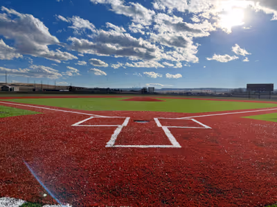 View from behind home plate of a baseball field with bright red artificial turf and partly cloudy sky.