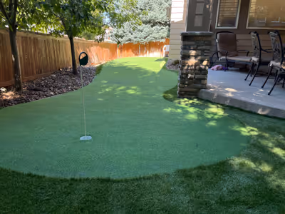 Backyard putting green with a hole and flag, bordered by a wooden fence, stone patio, and outdoor furniture.