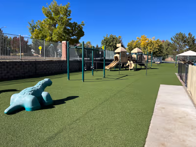 Playground with green artificial turf featuring a dinosaur-shaped climbing structure, swings, slides, and trees in the background under a clear blue sky.
