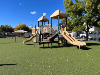 Playground structure with two beige slides, climbing elements, and green artificial turf under a clear blue sky.